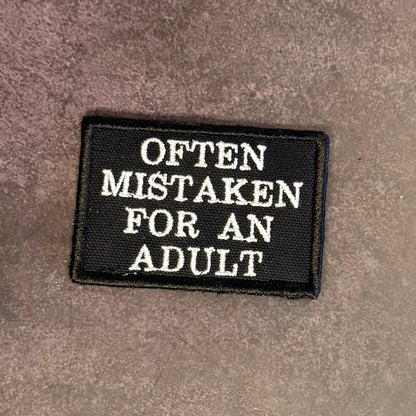 A rectangular black embroidered patch with white text reading 'OFTEN MISTAKEN FOR AN ADULT' on a textured gray background.