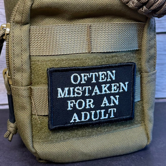 A close-up of a tactical pouch in olive drab/khaki fabric with a black rectangular patch that reads 'OFTEN MISTAKEN FOR AN ADULT' in white lettering. A braided paracord strap is wrapped around the top, and the pouch is photographed against a light wood plank background.