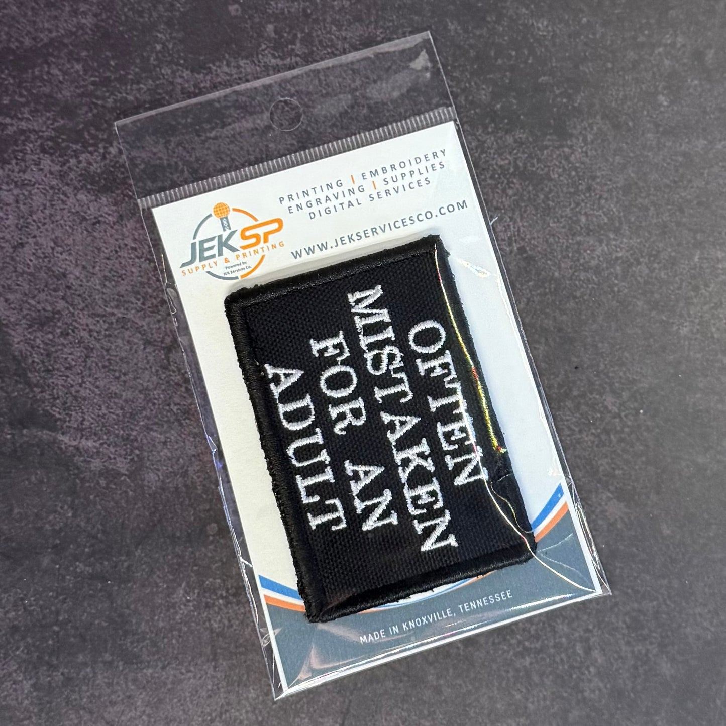 A black embroidered patch with white text reading 'OFTEN MISTAKEN FOR AN ADULT' inside a clear plastic retail package on a dark gray textured surface.
