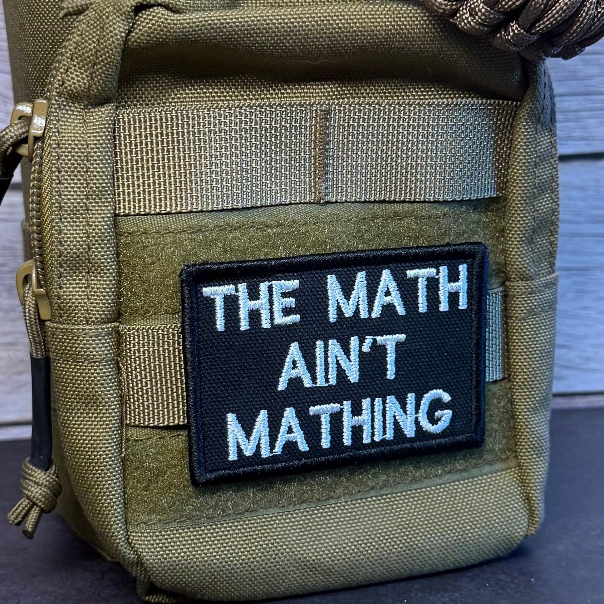 An olive green tactical pouch with a black embroidered patch that reads 'THE MATH AIN'T MATHING' in white text, shown against a rustic wood background.