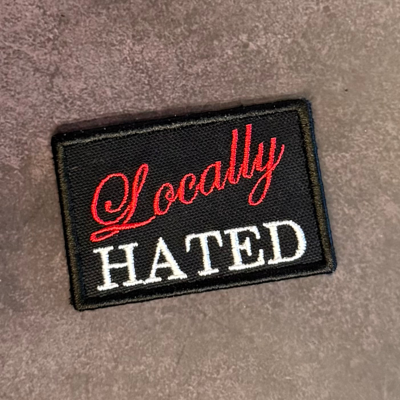 A rectangular black embroidered patch with the words 'Locally HATED' in red and white lettering, placed on a textured gray surface.