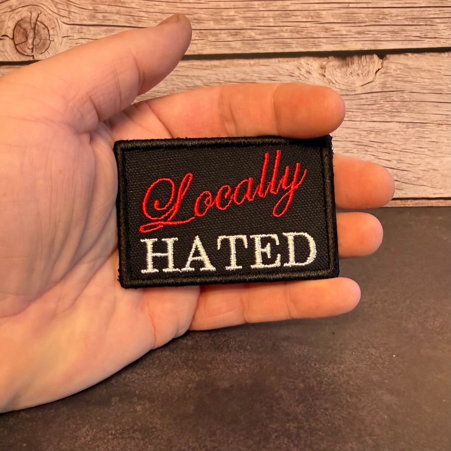 A hand holding a small rectangular black embroidered patch with the words 'Locally HATED' in red and white lettering, set against a rustic wood plank background.