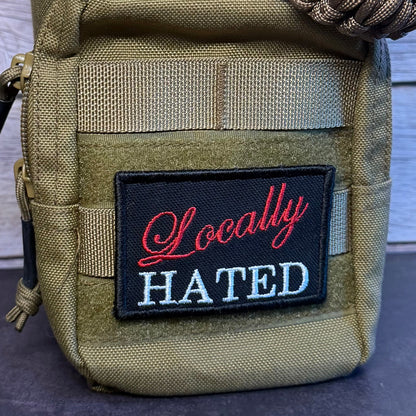 A tactical pouch in olive drab/khaki nylon with a black rectangular patch reading 'Locally HATED' in red and white lettering. A thick braided paracord strap is wrapped around the top, and the pouch is photographed against a rustic wood plank background.
