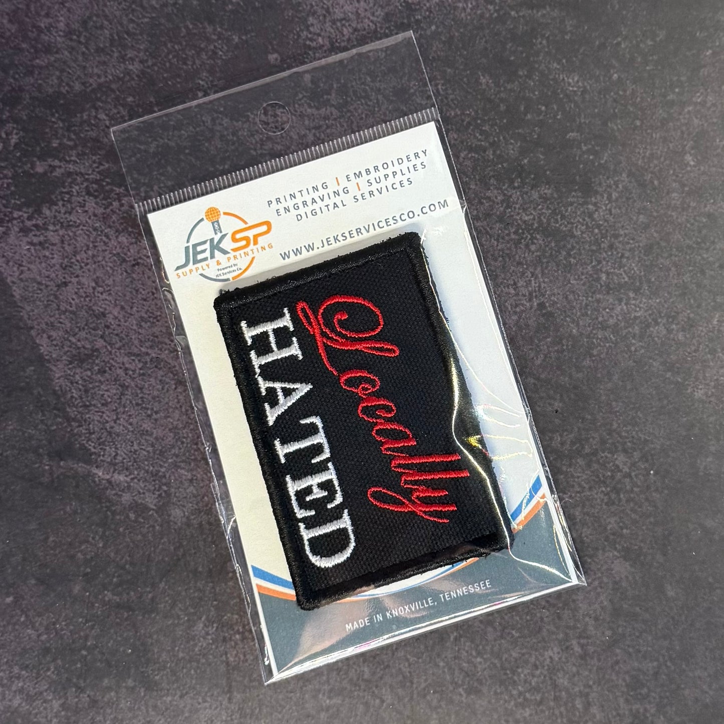 A black embroidered patch with white and red text reading 'HATE All of You' inside a clear plastic retail sleeve, photographed on a dark textured surface. The header card shows the brand JEKSP.