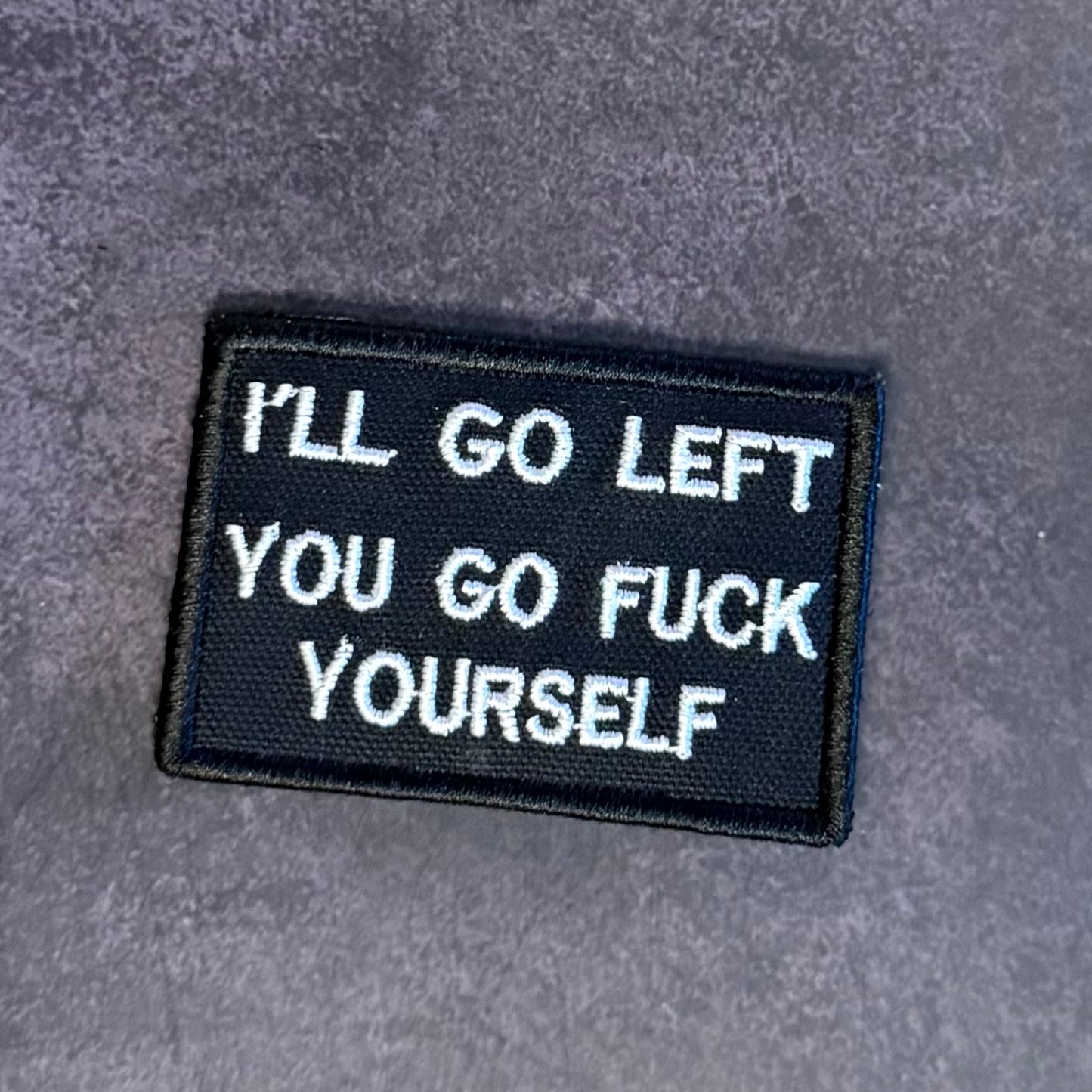 A rectangular black embroidered patch with white text reading 'I'LL GO LEFT YOU GO FUCK YOURSELF' on a textured gray background.
