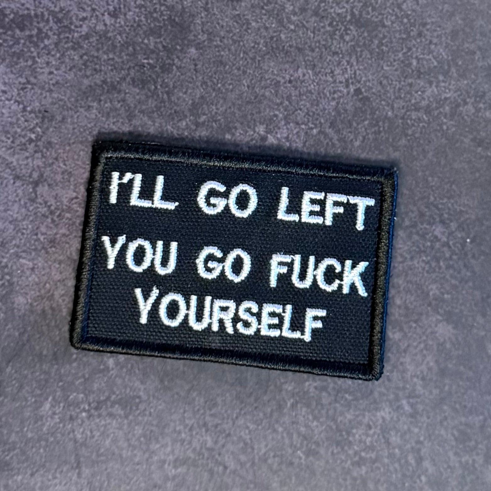 A rectangular black embroidered patch with white text reading 'I'LL GO LEFT YOU GO FUCK YOURSELF' on a textured gray background.