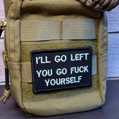 A tan tactical pouch with a black embroidered patch that reads 'I'LL GO LEFT YOU GO FUCK YOURSELF' in white text. The pouch has a woven strap across the top and is photographed against a rustic wood plank background.
