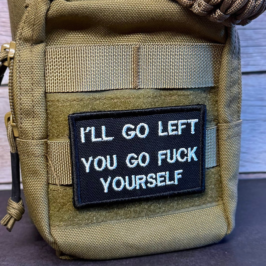 A tan tactical pouch with a black embroidered patch that reads 'I'LL GO LEFT YOU GO FUCK YOURSELF' in white text. The pouch has a woven strap across the top and is photographed against a rustic wood plank background.