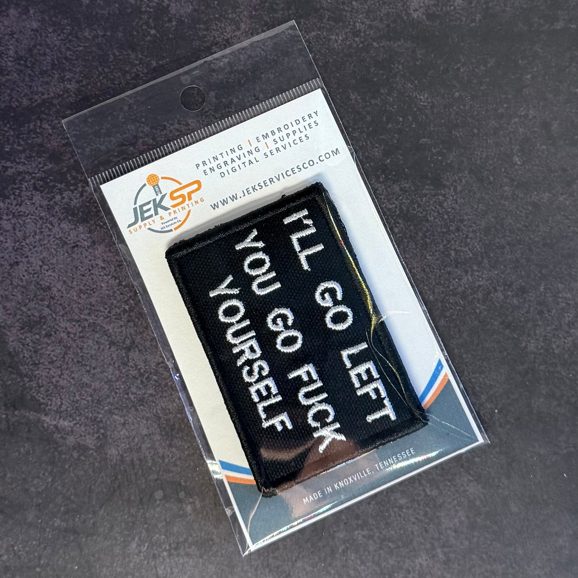 A black rectangular embroidered patch with white text reading 'I'LL GO LEFT YOU GO FUCK YOURSELF' in a clear plastic retail sleeve on a dark textured surface.