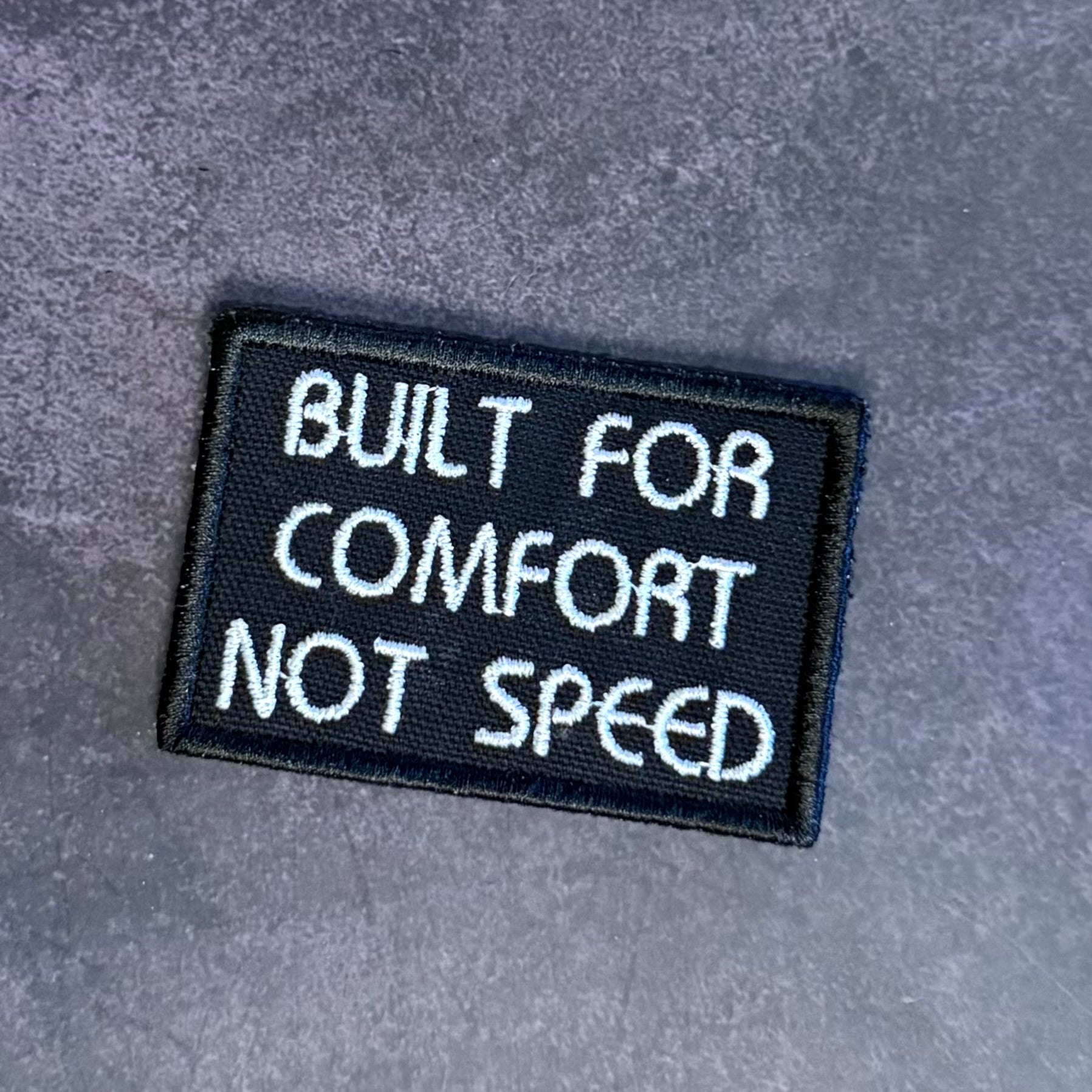 A rectangular embroidered patch with the text 'BUILT FOR COMFORT NOT SPEED' in white lettering on a black background, placed on a textured gray surface.