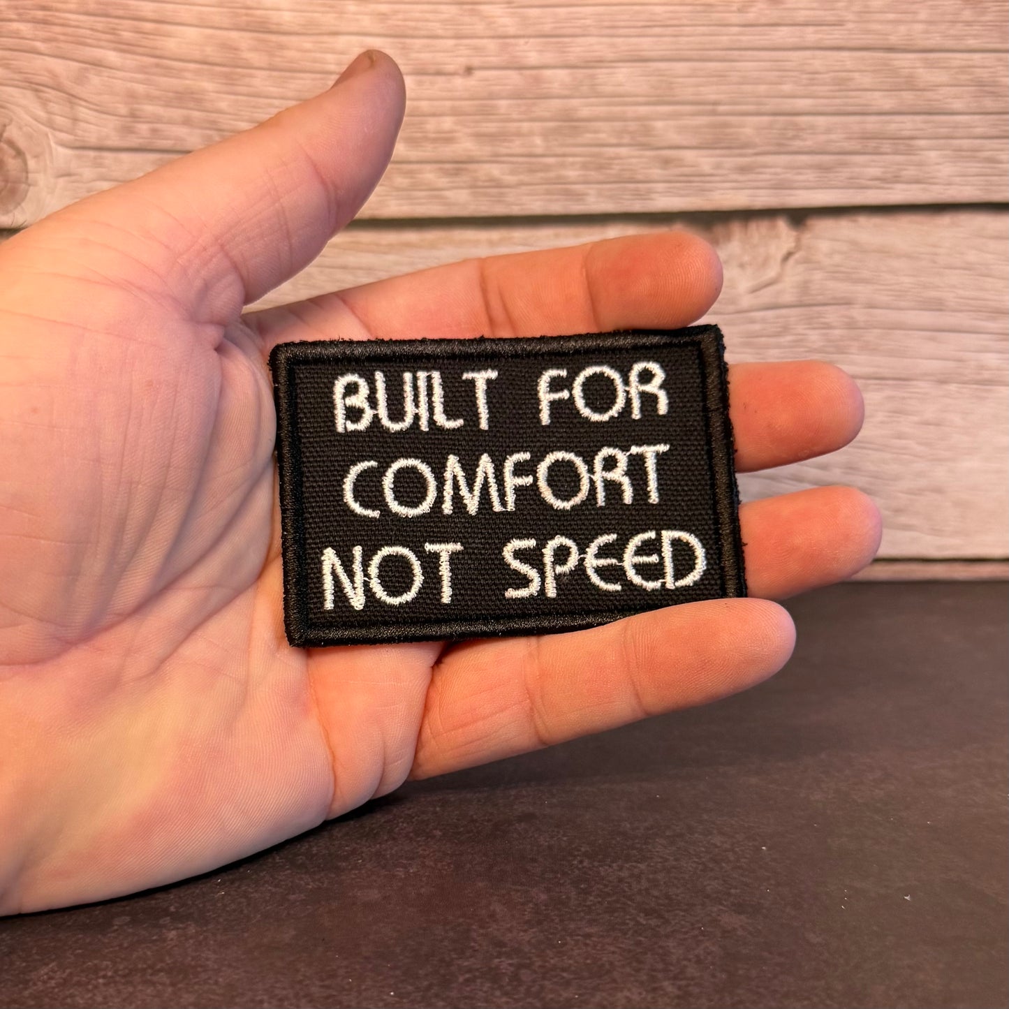 A hand holding a small rectangular black embroidered patch with white text that reads 'BUILT FOR COMFORT NOT SPEED' against a rustic wood and dark tabletop background.