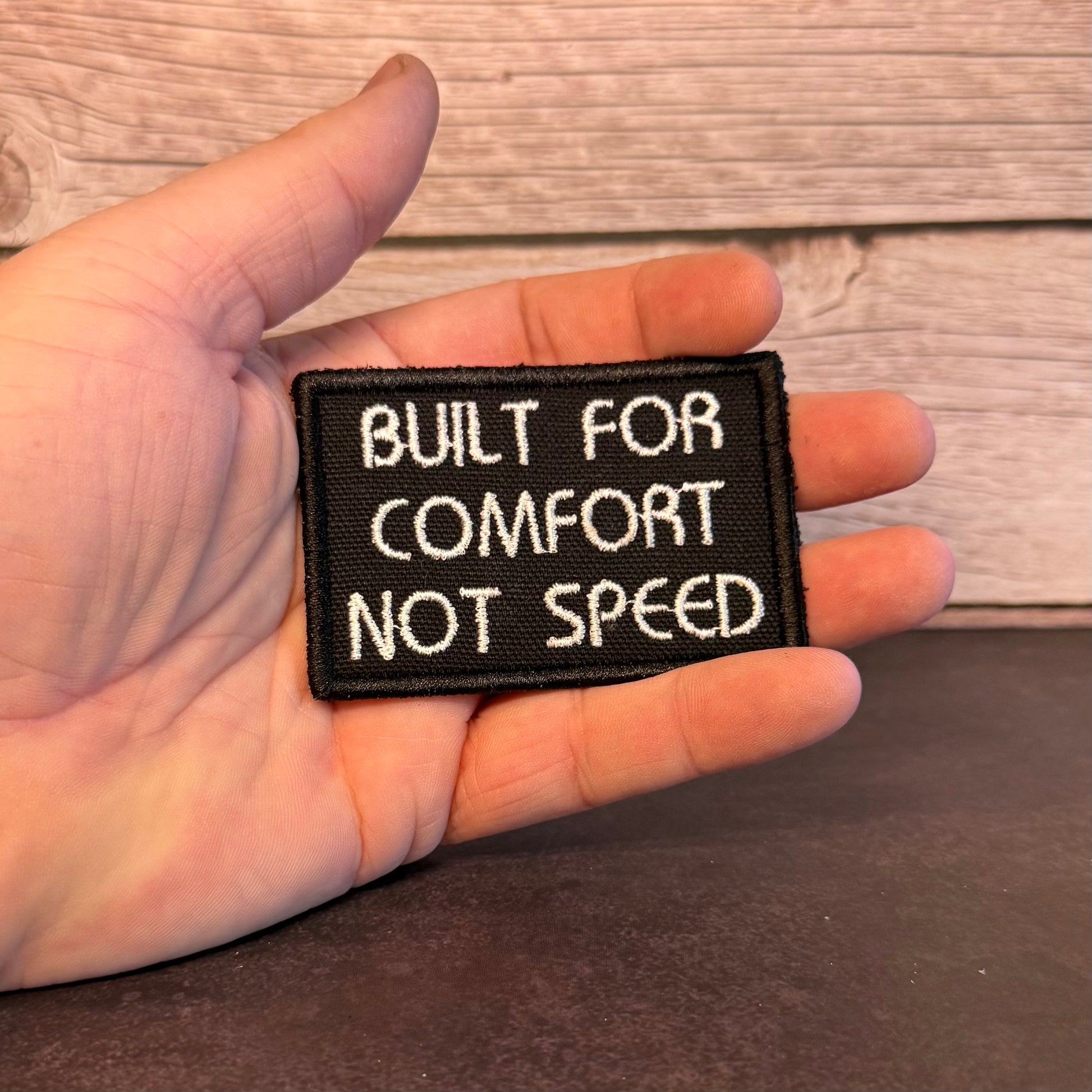 A hand holding a small rectangular black embroidered patch with white text that reads 'BUILT FOR COMFORT NOT SPEED' against a rustic wood and dark tabletop background.