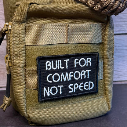 An olive drab tactical pouch with a black and white embroidered patch reading 'BUILT FOR COMFORT NOT SPEED' and a thick woven tan strap draped across the top, set against a rustic wood plank background.