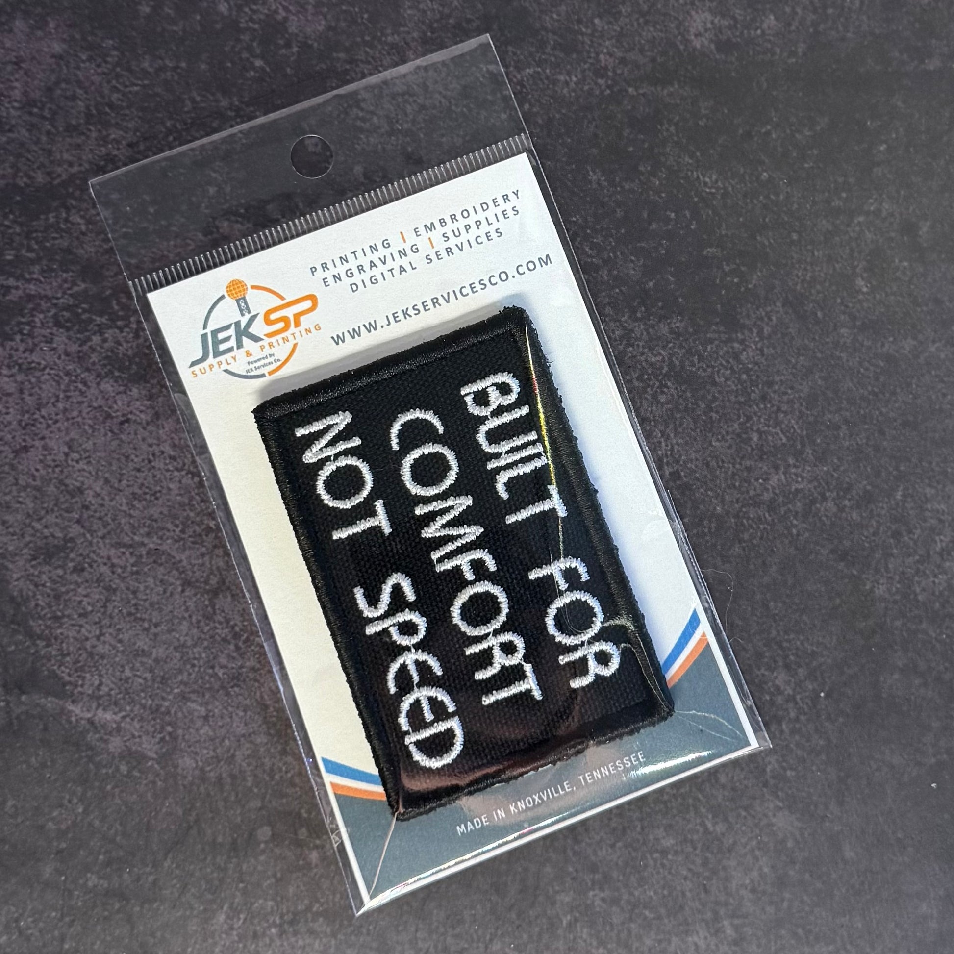 A black rectangular embroidered patch with white text reading 'BUILT FOR COMFORT NOT SPEED' inside a clear plastic retail sleeve with a branded backing card, placed on a dark textured surface.