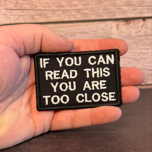 A hand holding a small rectangular black embroidered patch with white text that reads, 'IF YOU CAN READ THIS YOU ARE TOO CLOSE,' against a rustic wood and dark brown background.