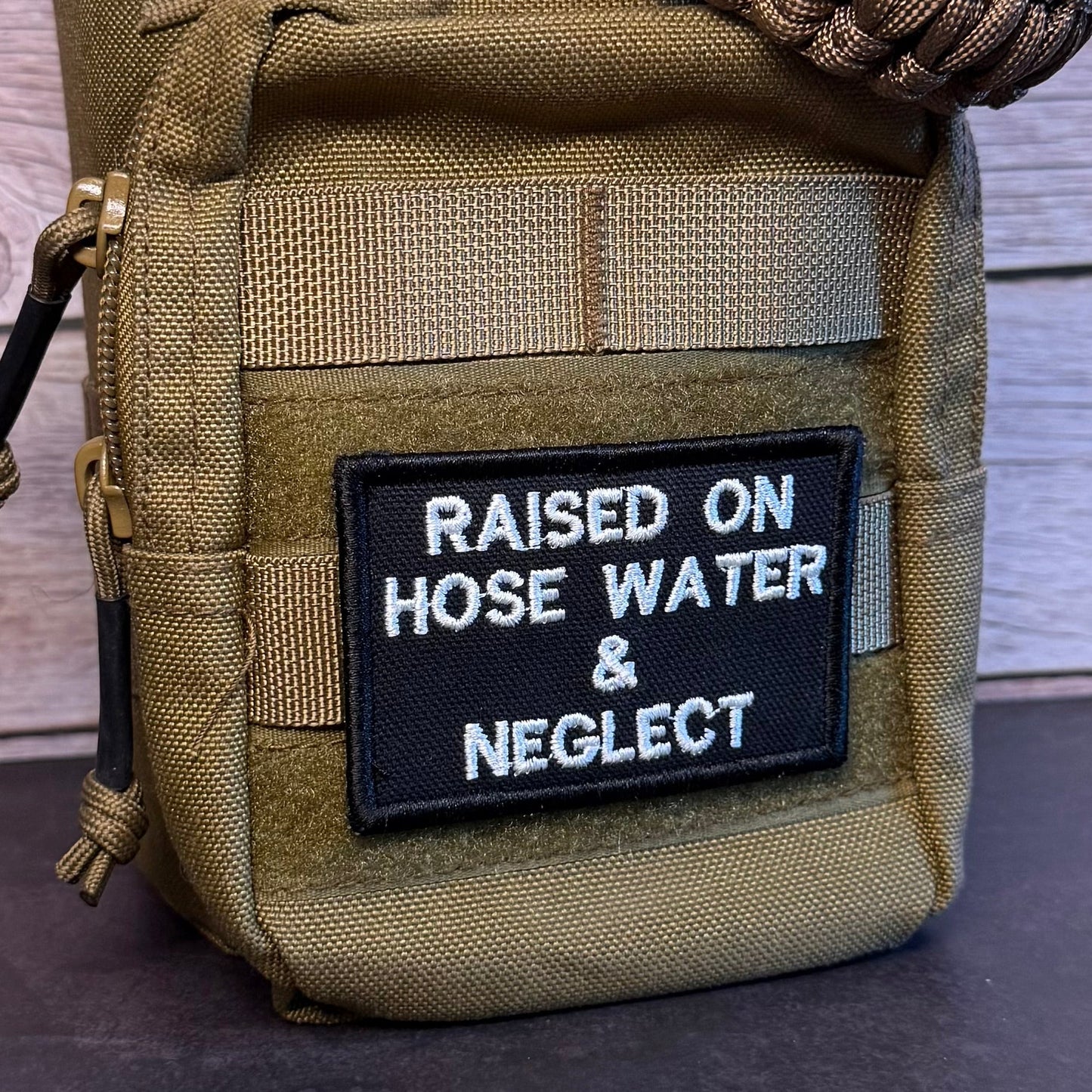 A close-up of a tactical utility pouch in olive drab/camouflage fabric, featuring a black rectangular patch with white embroidered text that reads 'RAISED ON HOSE WATER & NEGLECT'. A brown paracord strap is wrapped around the top portion of the pouch.