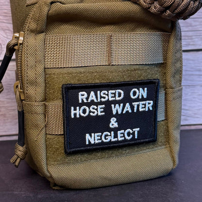 A close-up of a tactical utility pouch in olive drab/camouflage fabric, featuring a black rectangular patch with white embroidered text that reads 'RAISED ON HOSE WATER & NEGLECT'. A brown paracord strap is wrapped around the top portion of the pouch.