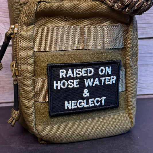 A close-up of a tactical utility pouch in olive drab/camouflage fabric, featuring a black rectangular patch with white embroidered text that reads 'RAISED ON HOSE WATER & NEGLECT'. A brown paracord strap is wrapped around the top portion of the pouch.