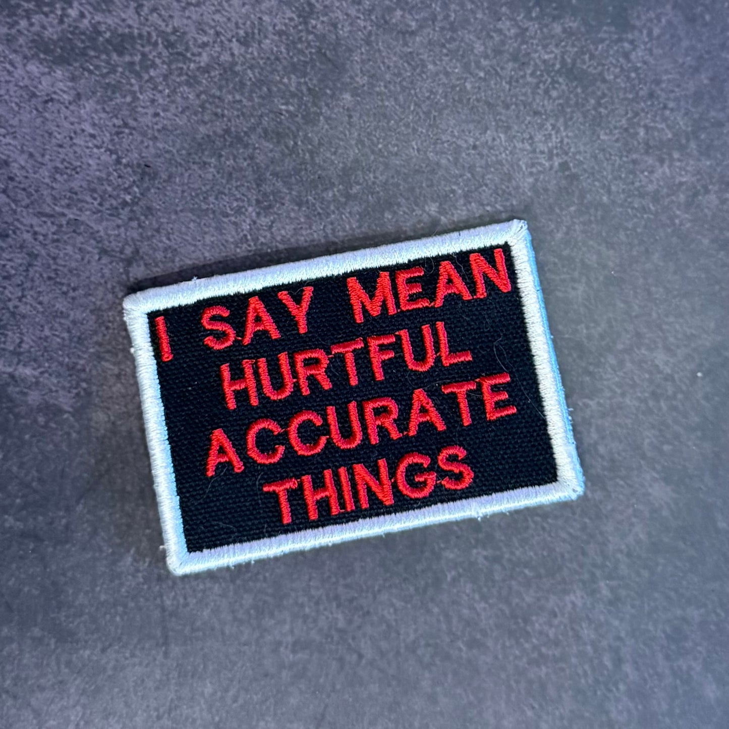 A rectangular embroidered patch with a black background, red text reading 'I SAY MEAN HURTFUL ACCURATE THINGS,' and a white border, placed on a dark gray textured surface.