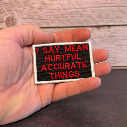 A hand holding a rectangular embroidered patch with red text reading 'I SAY MEAN HURTFUL ACCURATE THINGS' on a black background with a white border, against a rustic wood plank backdrop.