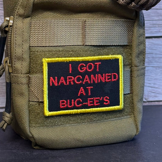 An olive drab tactical pouch with a black and red embroidered patch reading 'I GOT NARCANNED AT BUC-EE'S' in yellow border, shown with a paracord strap on a dark surface against a wood plank background.