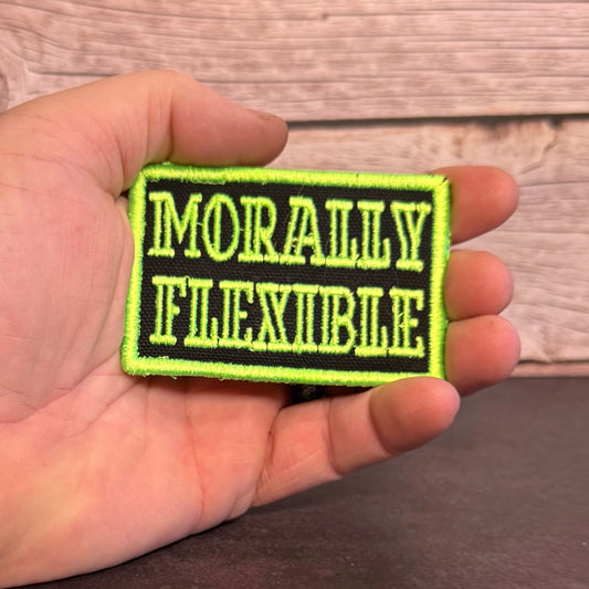 A hand holding a rectangular embroidered patch with the words 'MORALLY FLEXIBLE' in light green on a black background, with a bright neon green border, against a rustic wood plank backdrop.