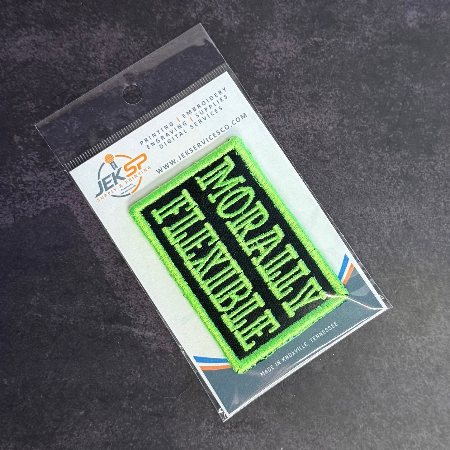 An embroidered rectangular patch reading 'MORALLY FLEXIBLE' in bright neon green on a black background, sealed in clear retail packaging with a printed header card. The product is photographed on a dark textured surface.