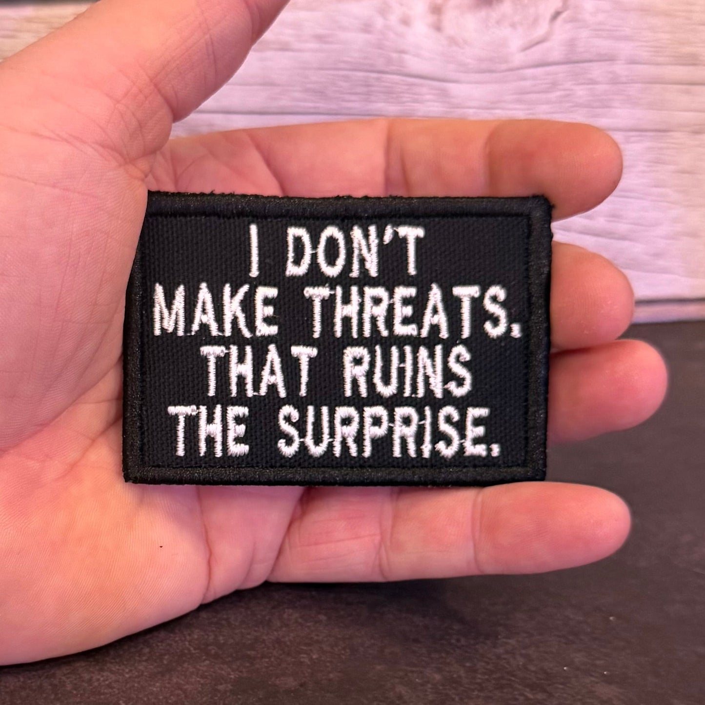 A hand holding a rectangular black embroidered patch with white text that reads, 'I DON'T MAKE THREATS. THAT RUINS THE SURPRISE.