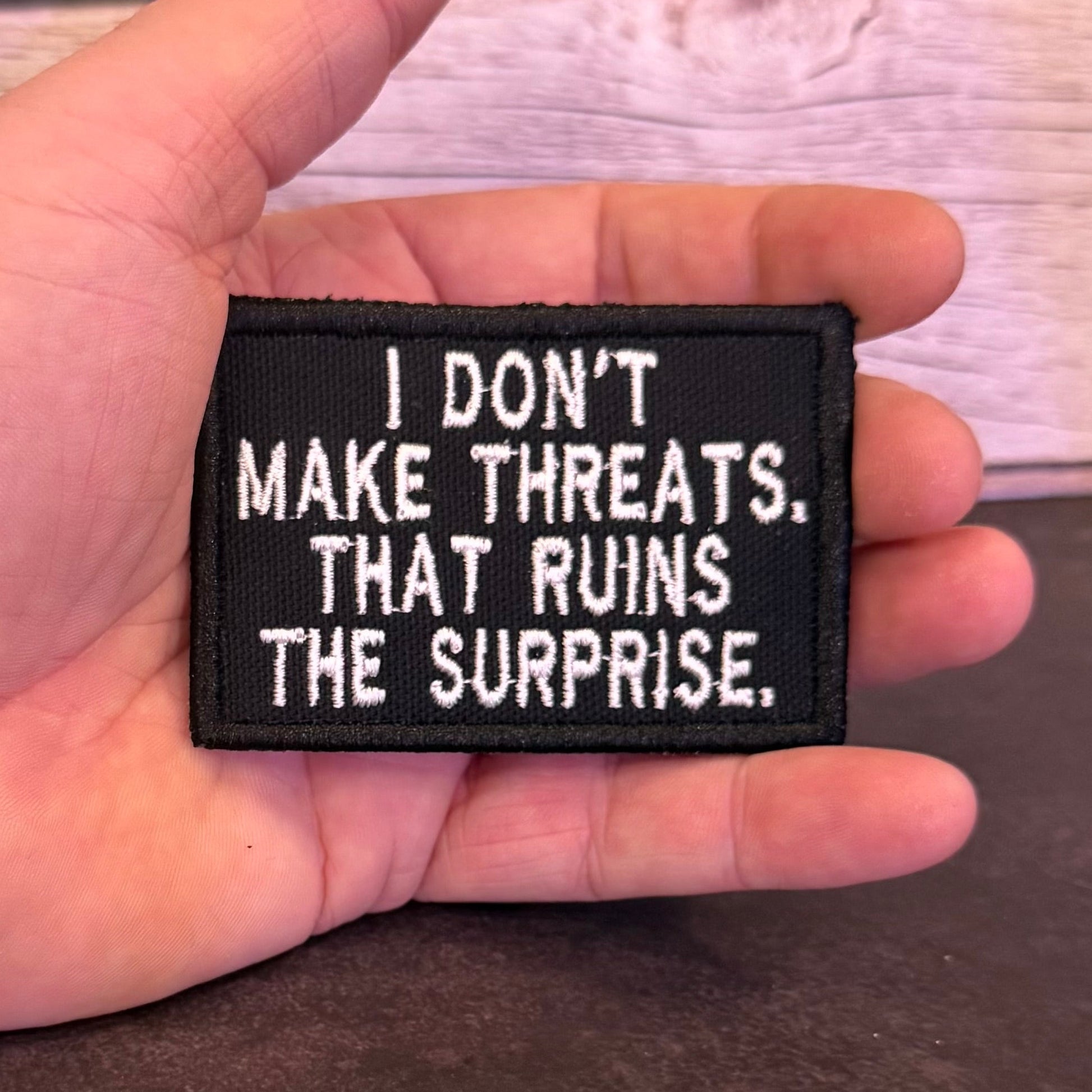 A hand holding a rectangular black embroidered patch with white text that reads, 'I DON'T MAKE THREATS. THAT RUINS THE SURPRISE.
