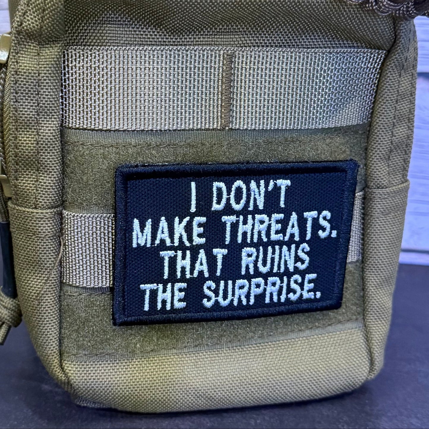 A tactical pouch in olive drab and coyote tan webbing, featuring a black rectangular patch with white embroidered text that reads, 'I DON'T MAKE THREATS. THAT RUINS THE SURPRISE.