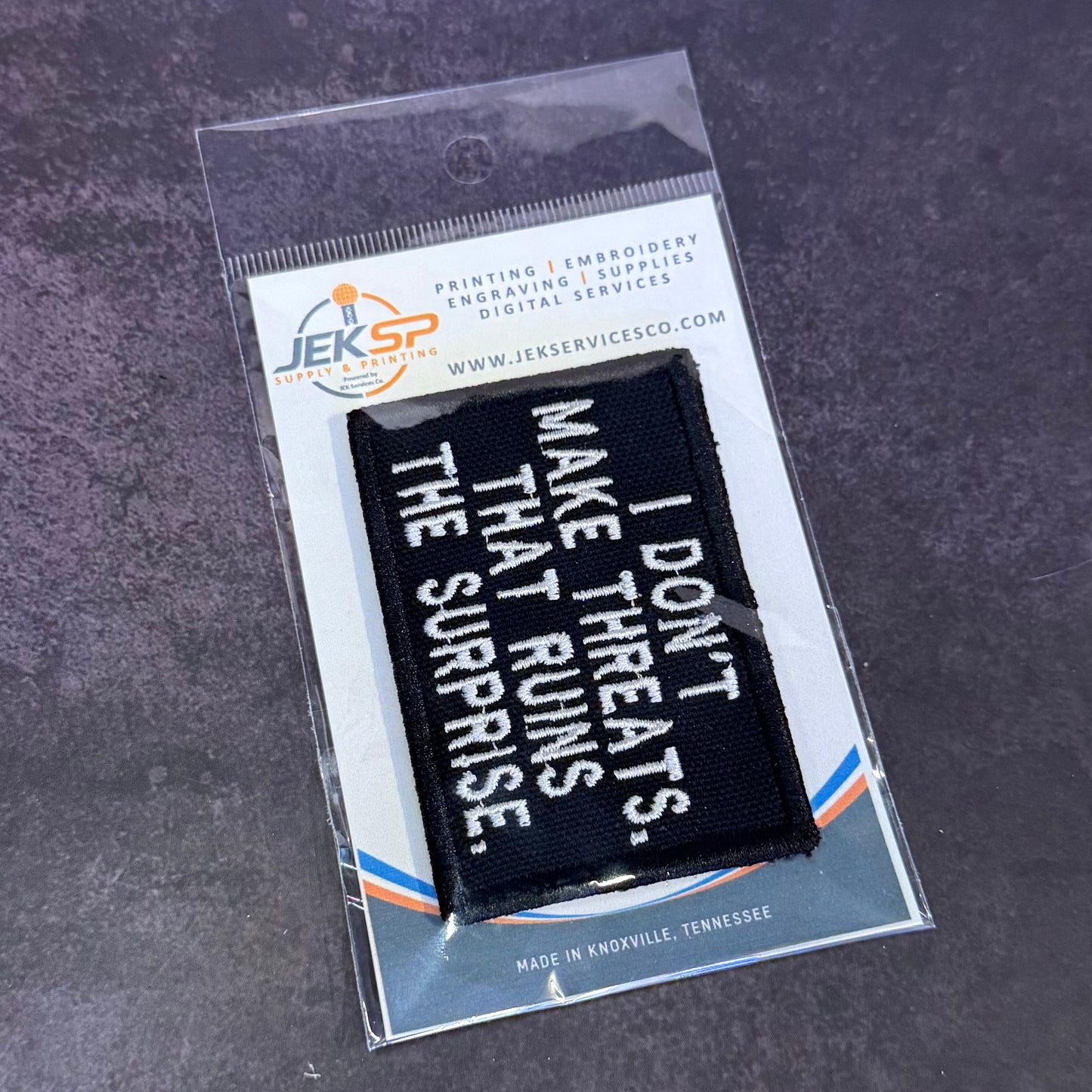 A black rectangular embroidered patch with white text reading 'I DON'T MAKE THREATS. THE SURPRISE.
