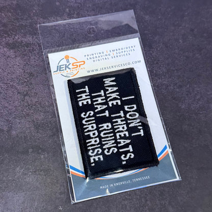 A black rectangular embroidered patch with white text reading 'I DON'T MAKE THREATS. THE SURPRISE.