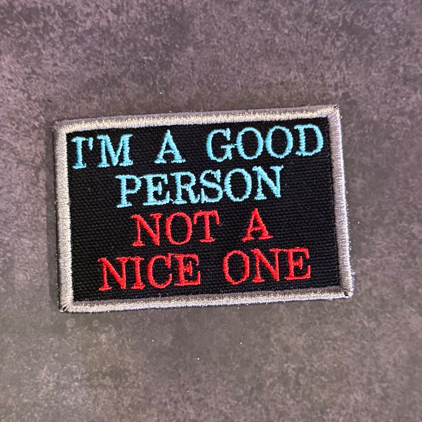 A rectangular embroidered patch with a black background and gray border, featuring the text 'I'M A GOOD PERSON NOT A NICE ONE' in light blue and red lettering, placed on a textured gray surface.