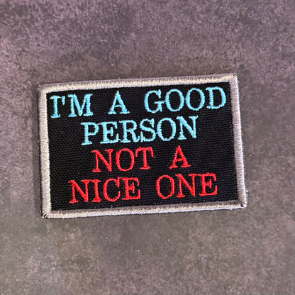 A rectangular embroidered patch with a black background and gray border, featuring the text 'I'M A GOOD PERSON NOT A NICE ONE' in light blue and red lettering, placed on a textured gray surface.