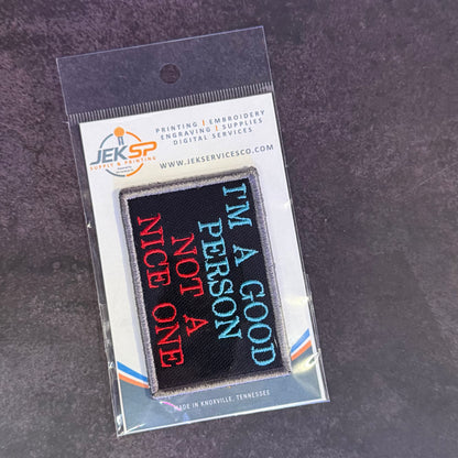 A rectangular embroidered patch in clear retail packaging, featuring the phrase “I’M A GOOD PERSON NOT A NICE ONE” in blue and red lettering on a black background. The header card shows the JKRSP logo and text for printing, embroidery, and digital services.