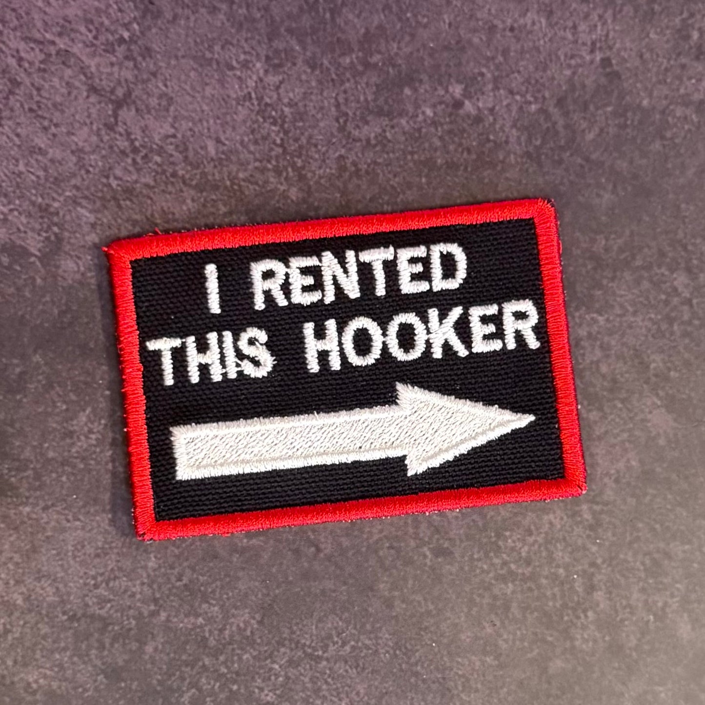 A rectangular embroidered patch with a black background, red border, white text reading 'I RENTED THIS HOOKER,' and a large white arrow pointing right, placed on a textured gray surface.