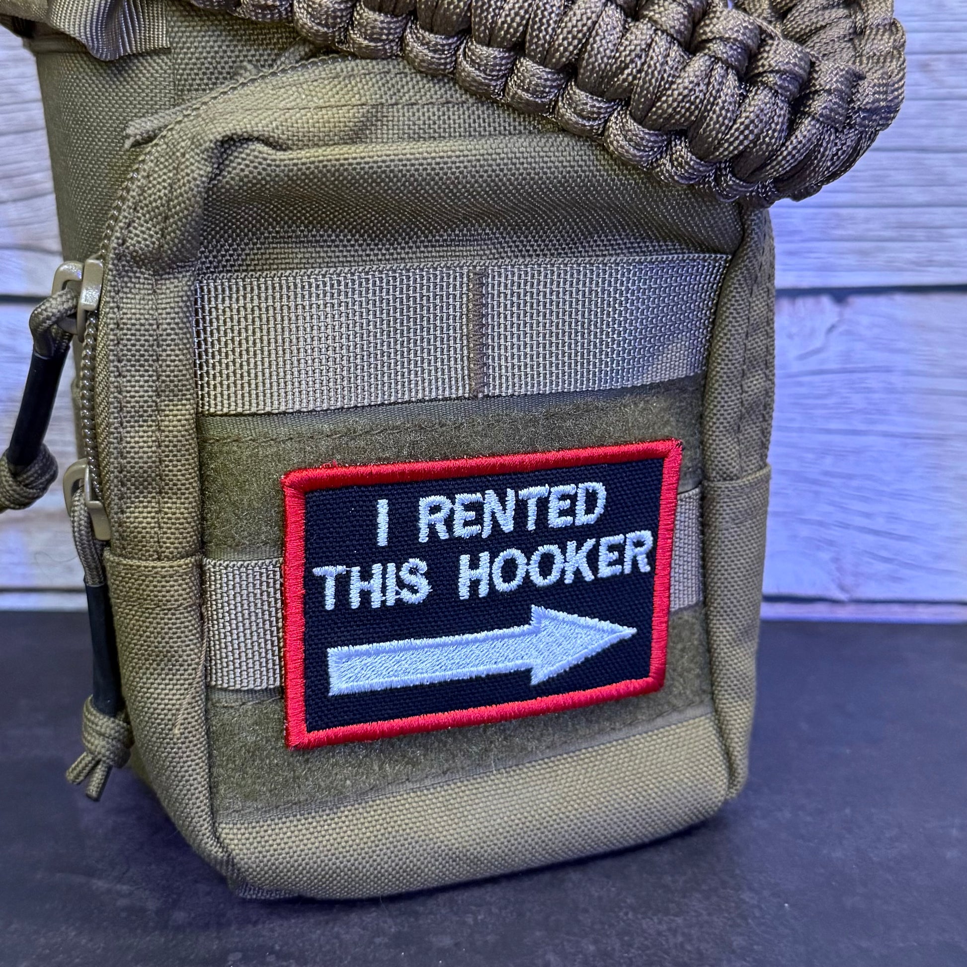 A close-up of a tan tactical pouch with a black embroidered patch that reads 'I RENTED THIS HOOKER' with a white arrow and red border. A woven strap and dark surface are visible in the frame.