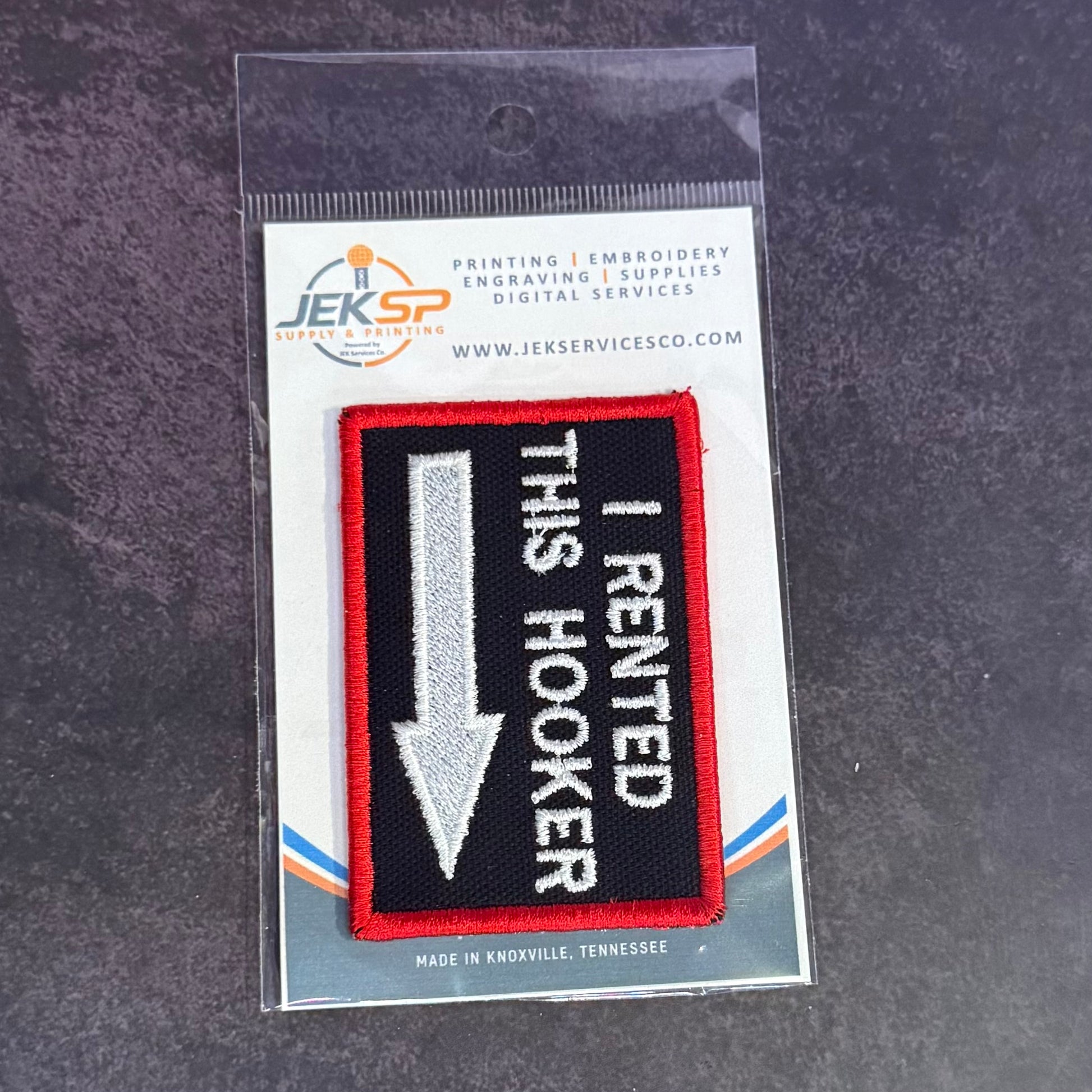 An embroidered patch in its original clear plastic packaging, featuring a black background with a red border, a silver arrow pointing down, and white text that reads 'I RENTED THIS HOOKER'. The packaging header card displays the brand 'Jeksp'.