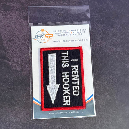 An embroidered patch in its original clear plastic packaging, featuring a black background with a red border, a silver arrow pointing down, and white text that reads 'I RENTED THIS HOOKER'. The packaging header card displays the brand 'Jeksp'.