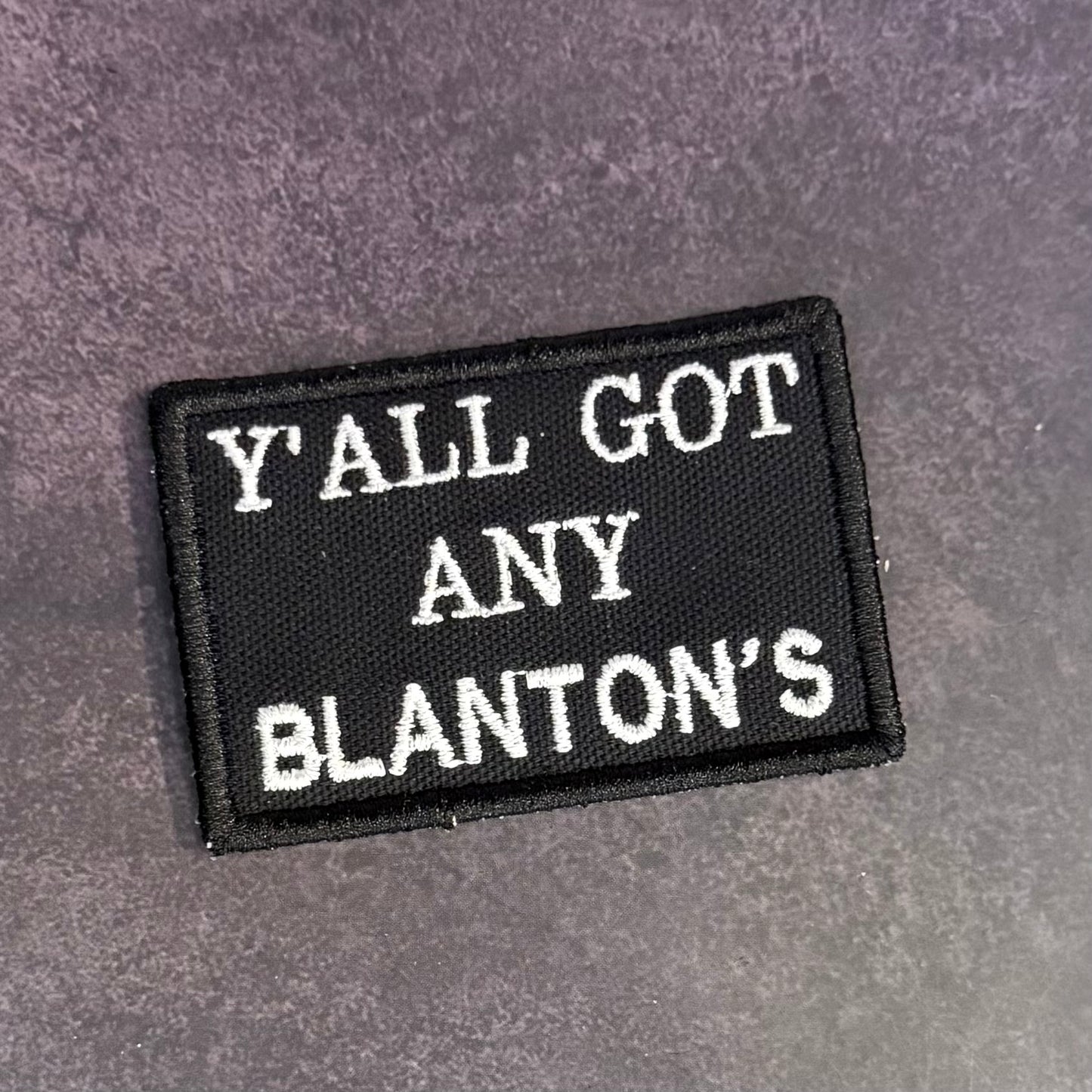 A rectangular black embroidered patch with white text reading 'Y'ALL GOT ANY BLANTON'S' on a textured gray background.