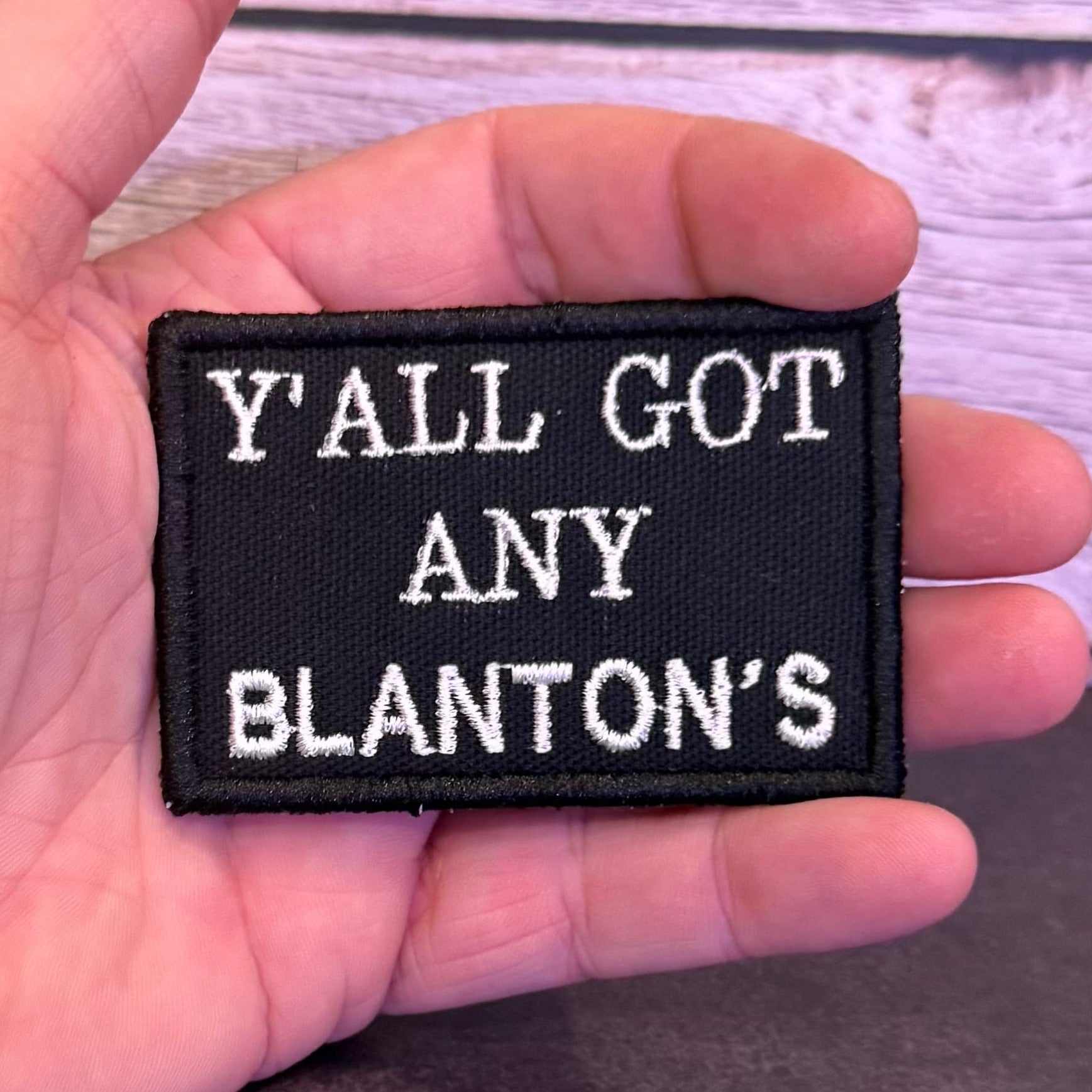 A hand holding a rectangular black embroidered patch with white text reading 'Y'ALL GOT ANY BLANTON'S' against a light wood plank background.