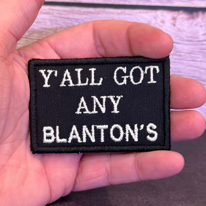 A hand holding a rectangular black embroidered patch with white text reading 'Y'ALL GOT ANY BLANTON'S' against a light wood plank background.