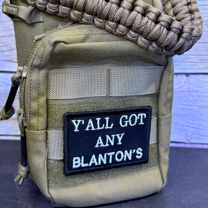 A close-up of a tactical pouch in olive drab/khaki nylon with a black embroidered patch that reads 'Y'ALL GOT ANY BLANTON'S' in white lettering. A tan paracord strap is threaded through the top, and the pouch is photographed against a light wood plank background.