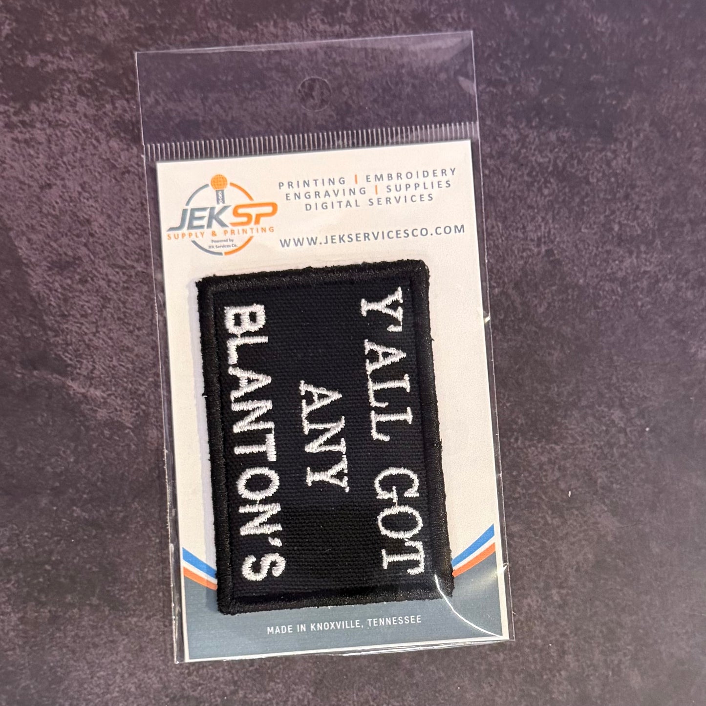 A black embroidered patch with white text reading 'Y'ALL GOT BLANTON'S ANY' displayed on a branded backing card inside a clear plastic sleeve, set against a dark textured background.