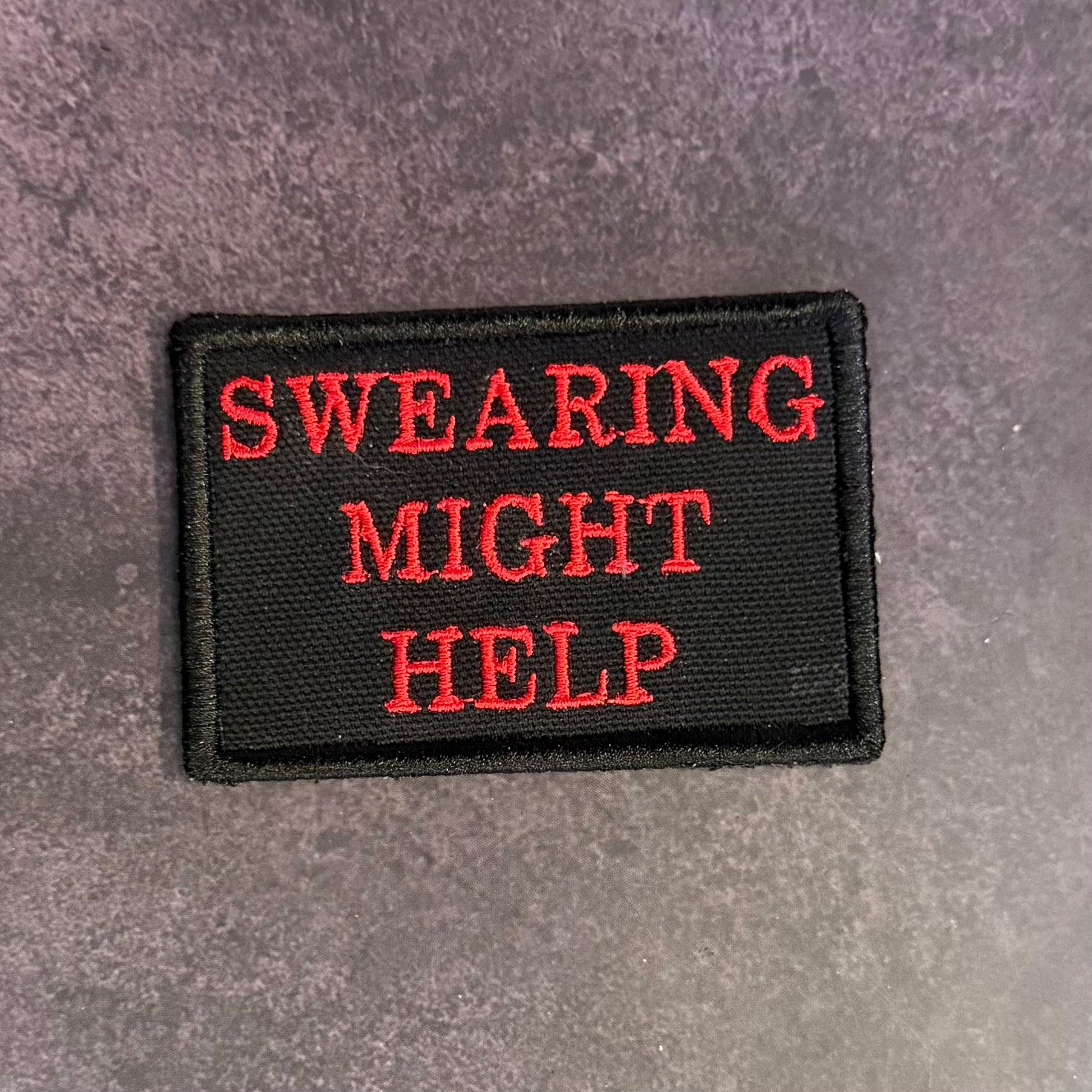 A rectangular black embroidered patch with the phrase 'SWEARING MIGHT HELP' in red capital letters, placed on a textured gray surface.