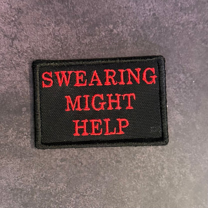 A rectangular black embroidered patch with the phrase 'SWEARING MIGHT HELP' in red capital letters, placed on a textured gray surface.