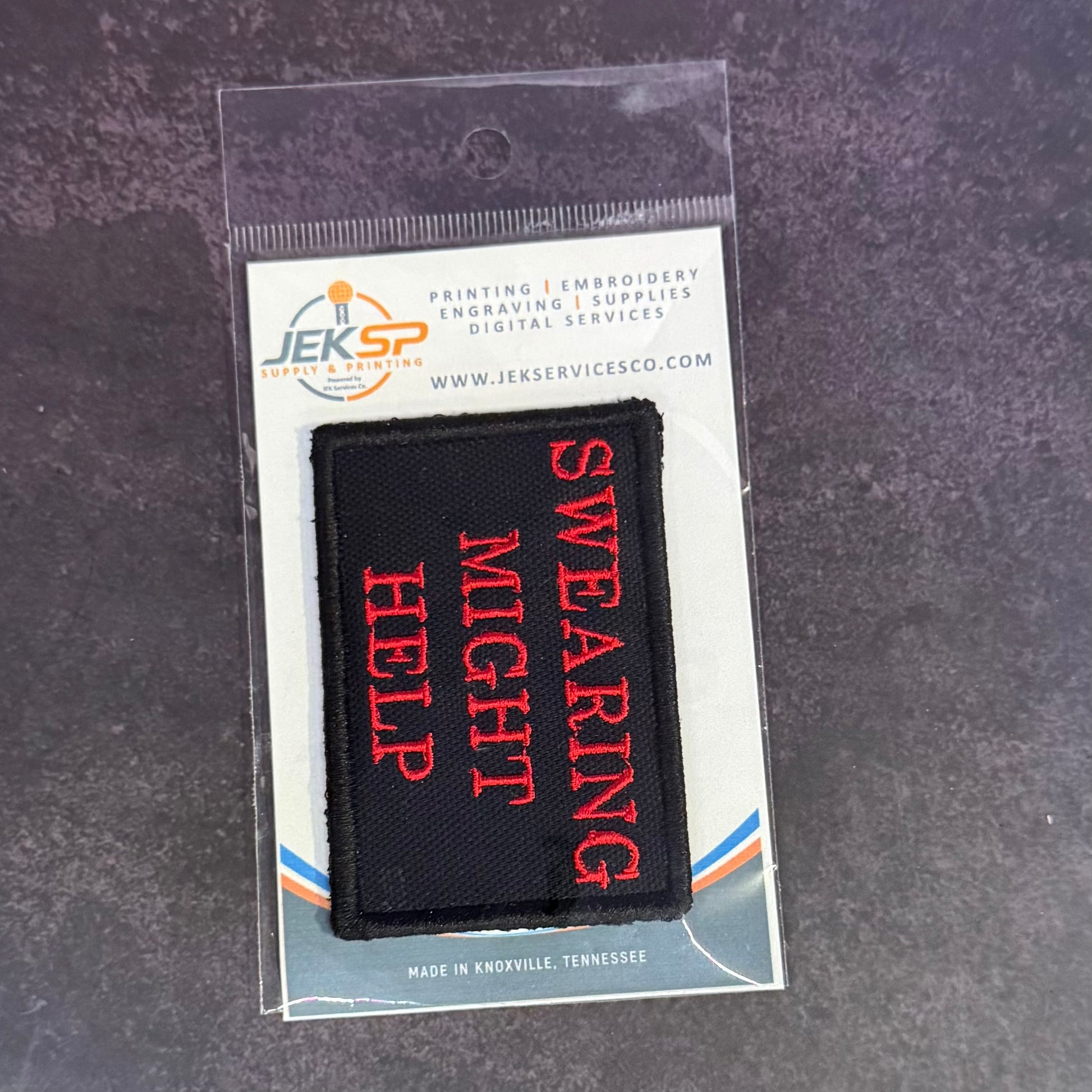 A black rectangular embroidered patch with red text reading 'SWEARING MIGHT HELP' inside a clear plastic hanging sleeve on a branded card, placed on a dark textured surface.
