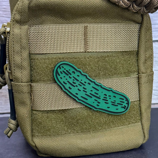 An olive green tactical pouch with webbing and a green pickle-shaped patch, shown close-up against a light wood background.