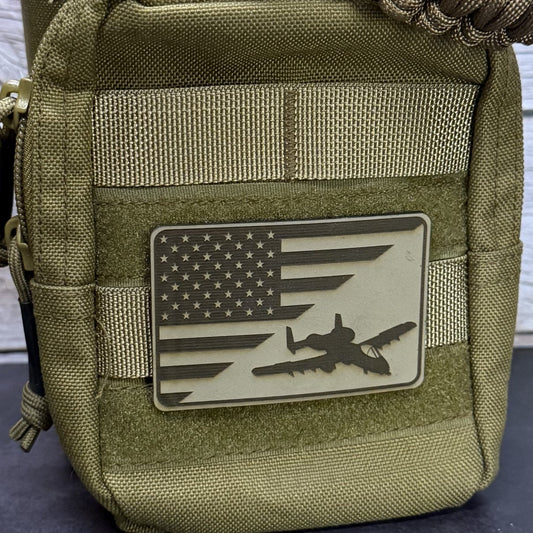 An olive drab tactical pouch featuring a rectangular PVC patch with a distressed American flag design and a black silhouette of an airplane, shown against a light wood background.