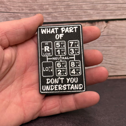 A hand holding a black rectangular patch with white text and car gear symbols. The patch reads, 'WHAT PART OF DON'T YOU UNDERSTAND.